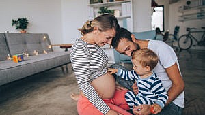 一张年轻的家庭坐在客厅地板上的照片。孩子摸着怀孕妈妈的肚子