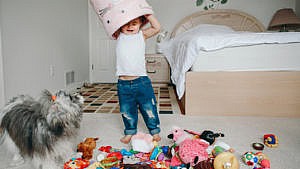 一个蹒跚学步的孩子把她的玩具扔掉，把粉红色的垃圾桶放在她的头上，而她的狗在旁边看着