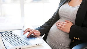 一位孕妇在她的笔记本电脑上工作，同时轻轻地把她的手放在肚子上。