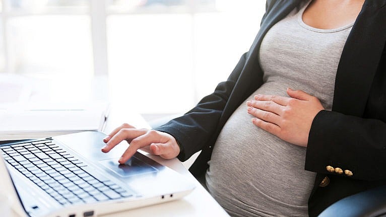 一位孕妇在她的笔记本电脑上工作，同时轻轻地把她的手放在肚子上。