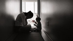 一张黑白照片，在狭窄的走廊里，一位母亲双手埋着头，而她的孩子坐在她身边。