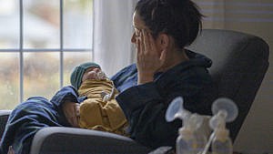 一位新妈妈穿着家常服坐在客厅的摇椅上，抱着刚出生的婴儿，身边有吸奶器和哺乳用品。