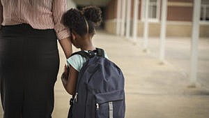 小学年龄，非裔美国女孩在上学前牵着妈妈或老师的手。她背着双肩包，抱着妈妈不确定是否要上学。