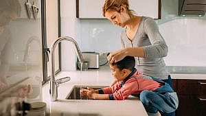 一个孩子在厨房的水槽边洗手，而他们的妈妈在一旁看着。