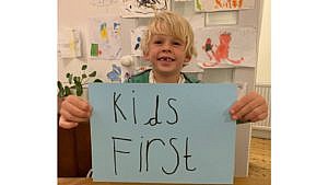 一个小孩在家庭娱乐室前举着手写的“孩子优先”的牌子，墙上挂着孩子们画的画